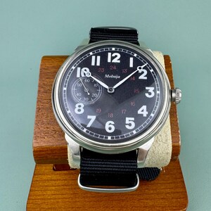 May include: A black-faced Molnija wristwatch with white numerals and a small seconds dial. The watch has a stainless steel case and a black fabric strap. The watch is displayed on a wooden stand.