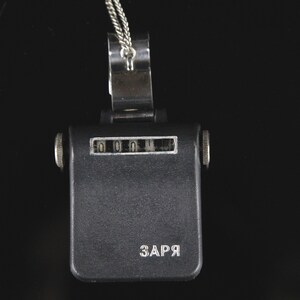 Pode incluir: Um contador preto com uma corrente prateada e um clipe prateado. O contador tem um visor que mostra "0000" e a palavra "ЗАРЯ" impressa na parte inferior.