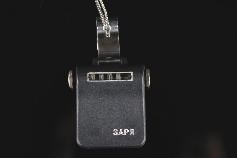 Pode incluir: Um contador preto com uma corrente prateada e um clipe prateado. O contador tem um visor que mostra "0000" e a palavra "ЗАРЯ" impressa na parte inferior.