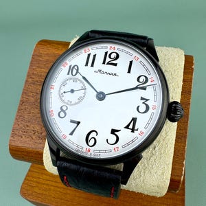 May include: A black wristwatch with a white face featuring large black numerals and red accents. The watch has a black leather band and a small seconds dial. The word "Molniya" is printed on the watch face.