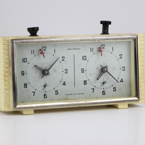 May include: A white vintage Jantar chess clock with two separate timers. The clock has a black knob on the top and is made in USSR.
