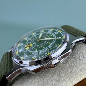 Puede incluir: Un reloj de pulsera vintage con esfera verde y caja plateada. El reloj tiene una correa de lona verde. La esfera del reloj presenta n&uacute;meros amarillos y un s&iacute;mbolo de radiaci&oacute;n.