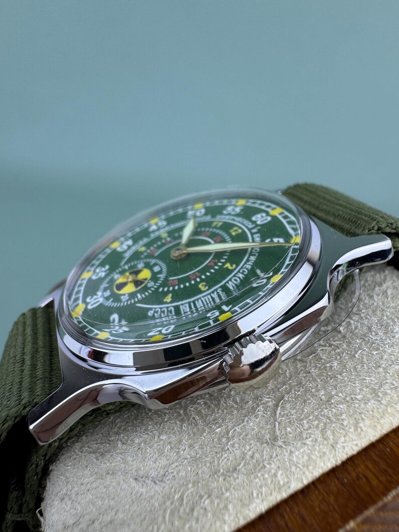Puede incluir: Un reloj de pulsera vintage con esfera verde y caja plateada. El reloj tiene una correa de lona verde. La esfera del reloj presenta n&uacute;meros amarillos y un s&iacute;mbolo de radiaci&oacute;n.