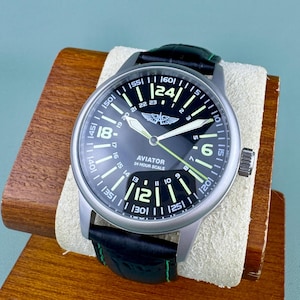 May include: A black Aviator watch with a stainless steel case and a black leather strap with green stitching. The watch face features white numerals, a 24-hour scale, and luminous hands. The watch is displayed on a wooden stand.