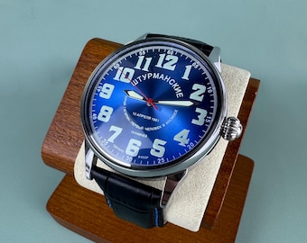 May include: A blue-faced wristwatch with white numerals and hands, and the text "ШТУРМАНСКИЕ". The watch has a black leather strap and a silver-coloured case. It is displayed on a wooden stand.