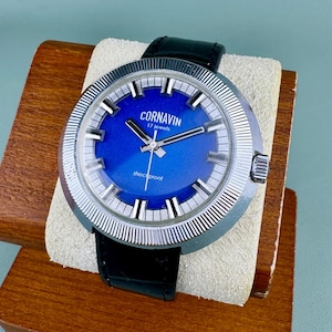 May include: A vintage Cornavin wristwatch with a bright blue face and silver bezel. The watch has a black leather strap and the text "Cornavin 17 jewels shockproof". The watch is displayed on a wooden stand.