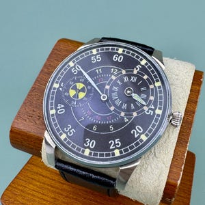 May include: A black wristwatch with a silver case and a black leather band. The watch face has white numerals and hands, with a smaller dial at the top. The watch is displayed on a wooden stand.