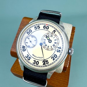 May include: A vintage wristwatch with a cream-colored face and black band. The watch features multiple sub-dials and the word "REGULATEUR". The watch is displayed on a wooden stand. The watch face has a diameter of approximately 40 mm.