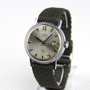 May include: Vintage silver-toned wristwatch with a green canvas band. The watch face displays the brand name, "CORNAVIN", and the text "17 JEWELS SHOCKPROOF". The watch has a date display at the 3 o'clock position and luminous hands.