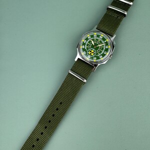 Puede incluir: Un reloj de pulsera con esfera verde y correa de nailon verde. La esfera del reloj tiene marcas blancas y amarillas, en una caja plateada. La correa se sujeta con una hebilla plateada.