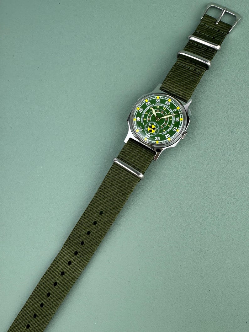 Puede incluir: Un reloj de pulsera con esfera verde y correa de nailon verde. La esfera del reloj tiene marcas blancas y amarillas, en una caja plateada. La correa se sujeta con una hebilla plateada.