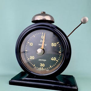 Puede incluir: Un temporizador negro vintage con una esfera redonda y una campana en la parte superior. El temporizador tiene una esfera verde oscuro con números dorados y la palabra "мин ты" en oro. El temporizador está sobre una base rectangular negra.