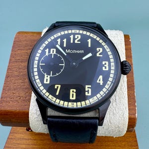 May include: A black wristwatch with a round face, featuring white numerals and hands against a black background. The watch has a leather strap and the word "Molniya" is printed on the face.