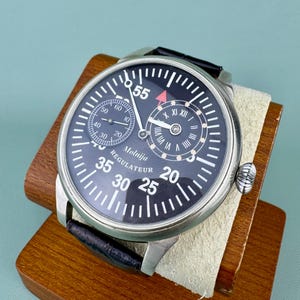May include: A black-faced Molnija Regulateur watch with a stainless steel case and black leather band. The watch features white numerals, a subdial, and a red-tipped second hand. The watch is displayed on a wooden stand.