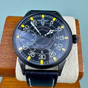 May include: Black wristwatch with a round face and a black leather band. The watch face has yellow and white numerals and markers. The watch is displayed on a wooden stand with a cream-colored cushion.