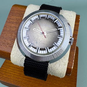 May include: A vintage wristwatch with a silver-colored bezel and a black fabric band. The watch face is gray with silver-colored hands and a red second hand. The watch is displayed on a wooden stand with a cream-colored cushion.