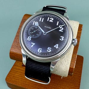May include: A black-faced Molnija wristwatch with white numerals and hands, and a small seconds dial. The watch has a stainless steel case and a black nylon strap. The watch is displayed on a wooden stand.