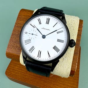 May include: A black wristwatch with a white face and black Roman numerals. The watch has a black leather band and is displayed on a wooden stand. The watch face has the word "Маяк" in cursive.