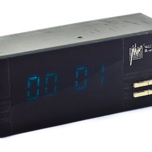 May include: A retro black digital clock showing "00 01" in blue digits. The clock has a rectangular form with buttons on the right and Cyrillic text. The design suggests a Soviet-era origin, with a focus on functionality.