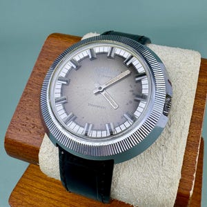 May include: A vintage Gorvana wristwatch with a silver-tone dial and hands, featuring the text "Gorvana 17 Jewels" and "Shockproof". The watch has a stainless steel case, a black leather band, and is displayed on a wooden stand.