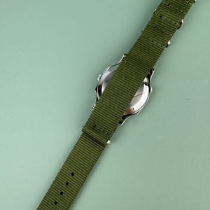 Puede incluir: Un reloj de pulsera plateado con una correa de nailon verde. El reloj tiene una esfera redonda y un cierre de hebilla. La correa mide aproximadamente 2 cm de ancho y tiene varios orificios de ajuste.