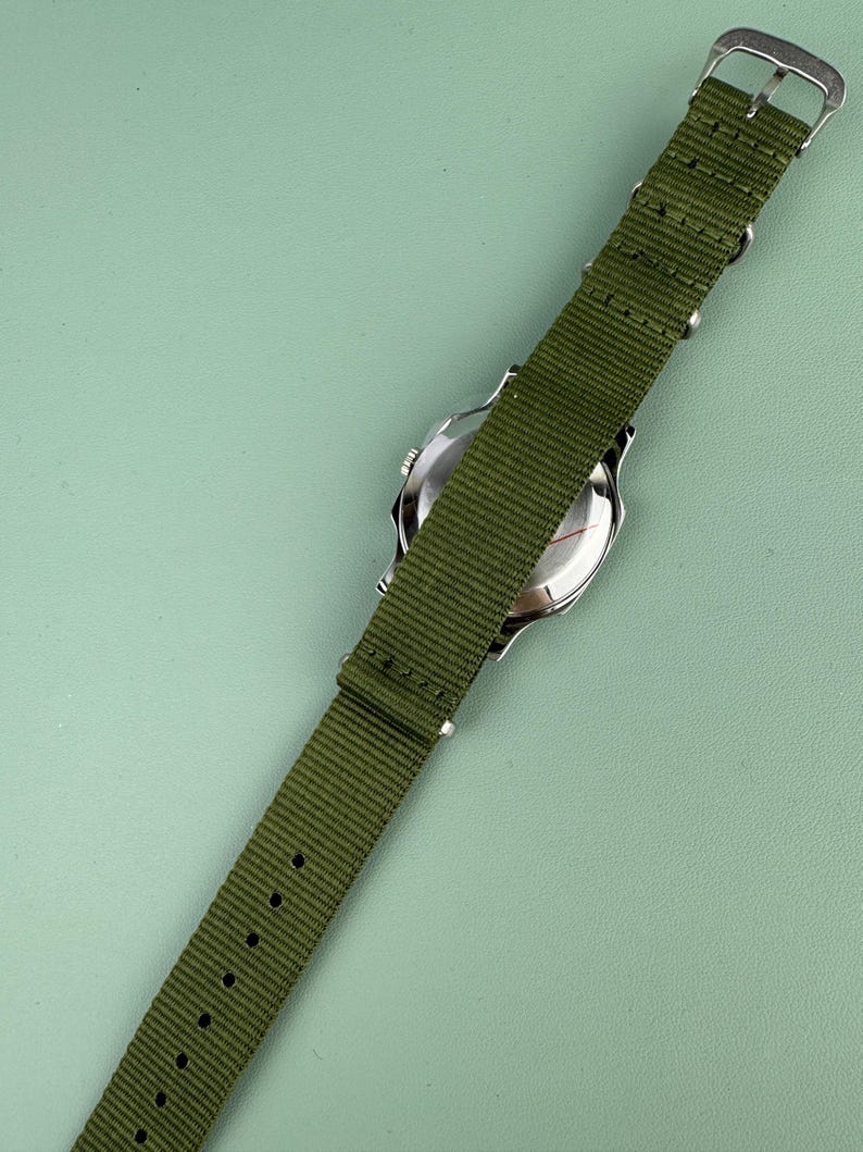 Puede incluir: Un reloj de pulsera plateado con una correa de nailon verde. El reloj tiene una esfera redonda y un cierre de hebilla. La correa mide aproximadamente 2 cm de ancho y tiene varios orificios de ajuste.