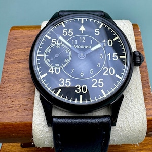 May include: A black Molnija wristwatch with a black leather band. The watch face is black with white numerals and hands. The watch is displayed on a wooden block.