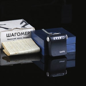 Pode incluir: Um ped&ocirc;metro preto com um visor digital e o texto "ЗАПР" no lado. O ped&ocirc;metro est&aacute; em uma caixa azul com o texto "ШАГОМЕР Пензенский ордена Ленина..." na parte superior de uma caixa branca.