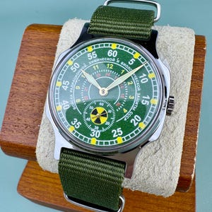 Puede incluir: Un reloj de pulsera verde y plateado con una correa de tela verde. La esfera del reloj es verde con n&uacute;meros blancos y un s&iacute;mbolo de peligro amarillo. El reloj se exhibe en un soporte de madera.