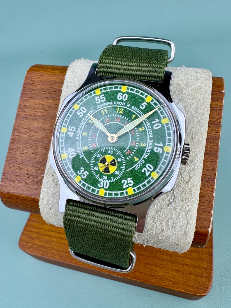 Puede incluir: Un reloj de pulsera verde y plateado con una correa de tela verde. La esfera del reloj es verde con n&uacute;meros blancos y un s&iacute;mbolo de peligro amarillo. El reloj se exhibe en un soporte de madera.