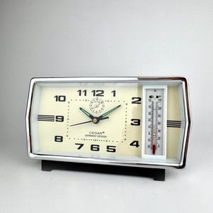 May include: A retro rectangular alarm clock with a cream face, black numerals, and a thermometer. The clock has a silver frame and the text "CEDAR GERMANY DESIGN". The thermometer displays temperatures in Celsius.