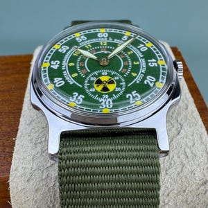 Puede incluir: Un reloj de pulsera vintage con esfera verde y caja de acero inoxidable. El reloj tiene una correa de lona verde y una esfera con marcas amarillas y blancas. La esfera del reloj tiene texto en cir&iacute;lico.