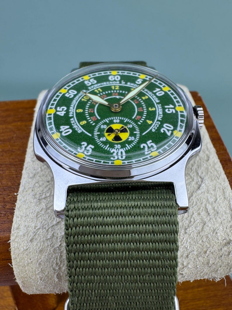 Puede incluir: Un reloj de pulsera vintage con esfera verde y caja de acero inoxidable. El reloj tiene una correa de lona verde y una esfera con marcas amarillas y blancas. La esfera del reloj tiene texto en cir&iacute;lico.