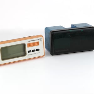 May include: Two vintage electronic devices. The first is a small, orange handheld device with a digital display and buttons. The second is a larger, black device with a blue top and a digital display. Both devices have the text "Time Technology" on them.