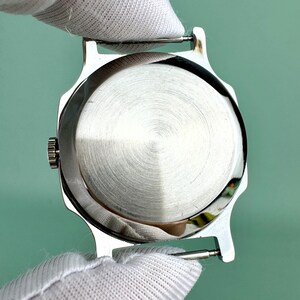 Puede incluir: Una caja de reloj plateada con una parte trasera de metal cepillado. La caja del reloj est&aacute; sujeta por una persona que lleva un guante blanco. El reloj tiene una peque&ntilde;a corona y asas para sujetar una correa.