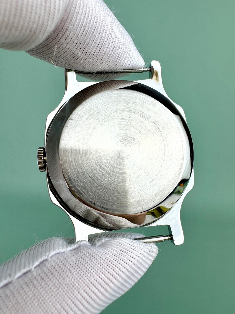 Puede incluir: Una caja de reloj plateada con una parte trasera de metal cepillado. La caja del reloj est&aacute; sujeta por una persona que lleva un guante blanco. El reloj tiene una peque&ntilde;a corona y asas para sujetar una correa.