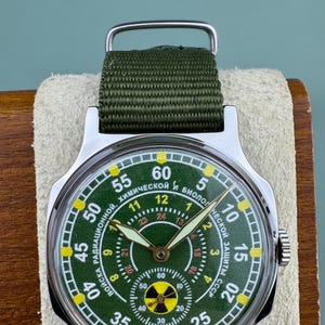 Puede incluir: Reloj de pulsera vintage con esfera verde y correa de tela verde. La esfera del reloj presenta n&uacute;meros blancos y amarillos y texto en cir&iacute;lico. El reloj tiene una caja de acero inoxidable y un dise&ntilde;o militar.
