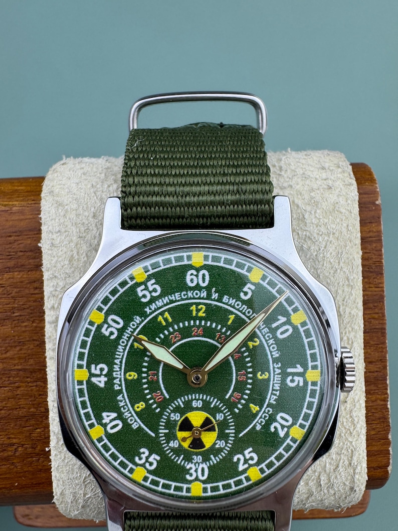 Puede incluir: Reloj de pulsera vintage con esfera verde y correa de tela verde. La esfera del reloj presenta n&uacute;meros blancos y amarillos y texto en cir&iacute;lico. El reloj tiene una caja de acero inoxidable y un dise&ntilde;o militar.