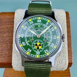 Puede incluir: Un reloj de estilo militar con esfera verde y correa de nailon verde. La esfera del reloj presenta un s&iacute;mbolo de radiaci&oacute;n y texto en idioma extranjero. El reloj se muestra sobre un soporte de madera.