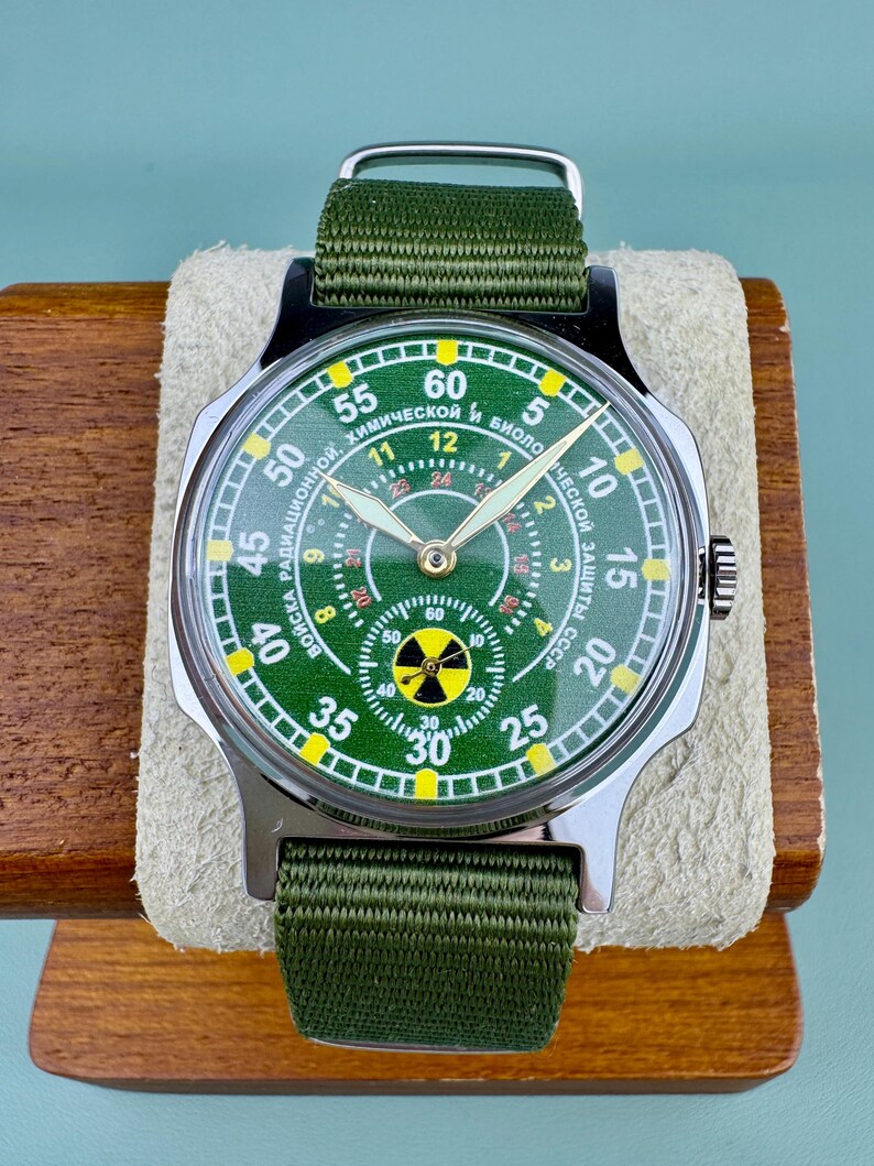 Puede incluir: Un reloj de estilo militar con esfera verde y correa de nailon verde. La esfera del reloj presenta un s&iacute;mbolo de radiaci&oacute;n y texto en idioma extranjero. El reloj se muestra sobre un soporte de madera.