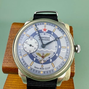 May include: A vintage wristwatch with a silver case and a black nylon strap. The watch face is light blue with a red star, a winged emblem, and the text "Заказ ВМФ СССР". The watch is displayed on a wooden stand.