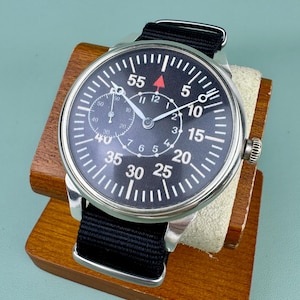 May include: A black-faced wristwatch with white numerals and a red triangle at the 12 o'clock position. The watch has a black nylon strap and a silver-colored case. It rests on a wooden display stand.