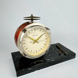 Op de afbeelding: Een vintage klok in de vorm van een trommel met een zilverkleurige rand en een crèmekleurige wijzerplaat. De klok heeft goudkleurige wijzers en het woord "Механика" op de wijzerplaat. Hij staat op een zwarte marmeren voet met drumstokken. De klok is rood.