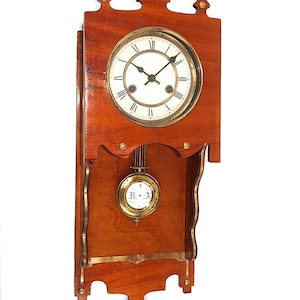 Könnte beinhalten: Eine Wanduhr aus Holz mit einem Messingzifferblatt und einem Pendel. Die Uhr hat ein Zifferblatt mit römischen Ziffern und einen weißen Hintergrund. Die Uhr befindet sich in einem Holzrahmen mit verzierten Schnitzereien.