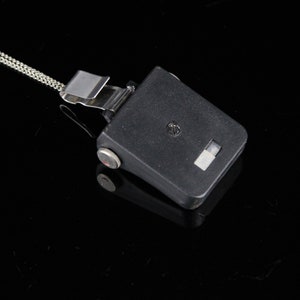 Pode incluir: Um dispositivo de pl&aacute;stico preto com um clipe prateado e uma corrente presa. O dispositivo tem um pequeno ret&acirc;ngulo branco na frente.