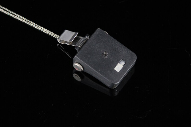 Pode incluir: Um dispositivo de pl&aacute;stico preto com um clipe prateado e uma corrente presa. O dispositivo tem um pequeno ret&acirc;ngulo branco na frente.