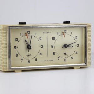 May include: A white analog chess clock with two separate faces. Each face has black hands and black numbers. The clock is labeled "Sandoz" and "Made in USSR".
