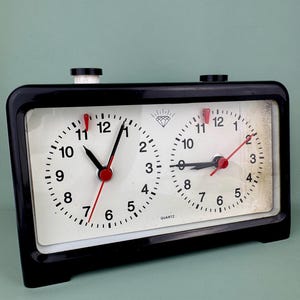 May include: A black quartz chess clock with two white clock faces. Each face has black numbers and minute markers, with red second hands and black hour and minute hands. The clock has two black buttons on top.
