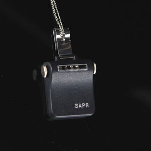 Pode incluir: Um pingente de metal preto com uma corrente de prata. O pingente tem uma forma retangular com um pequeno bot&atilde;o de prata de cada lado. O pingente tem a palavra "ЗАРЯ" inscrita na frente.