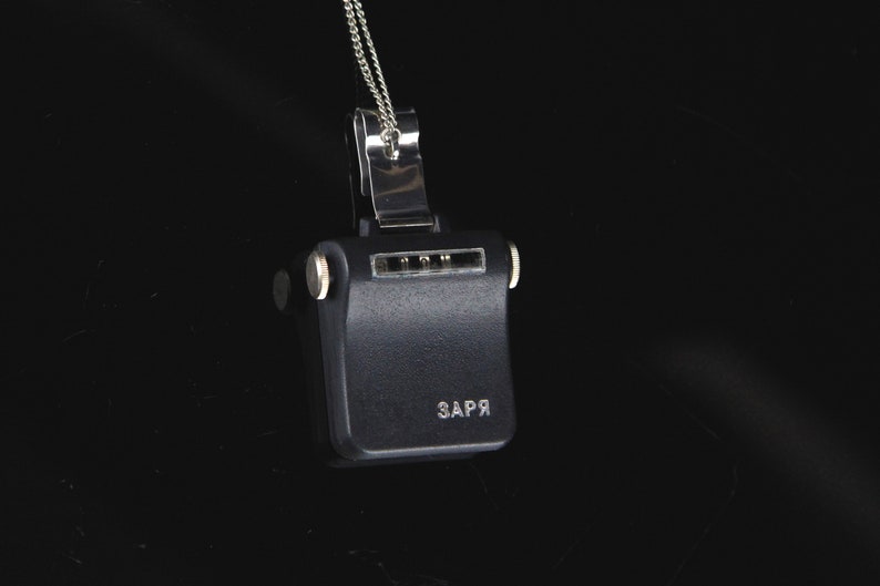 Pode incluir: Um pingente de metal preto com uma corrente de prata. O pingente tem uma forma retangular com um pequeno bot&atilde;o de prata de cada lado. O pingente tem a palavra "ЗАРЯ" inscrita na frente.